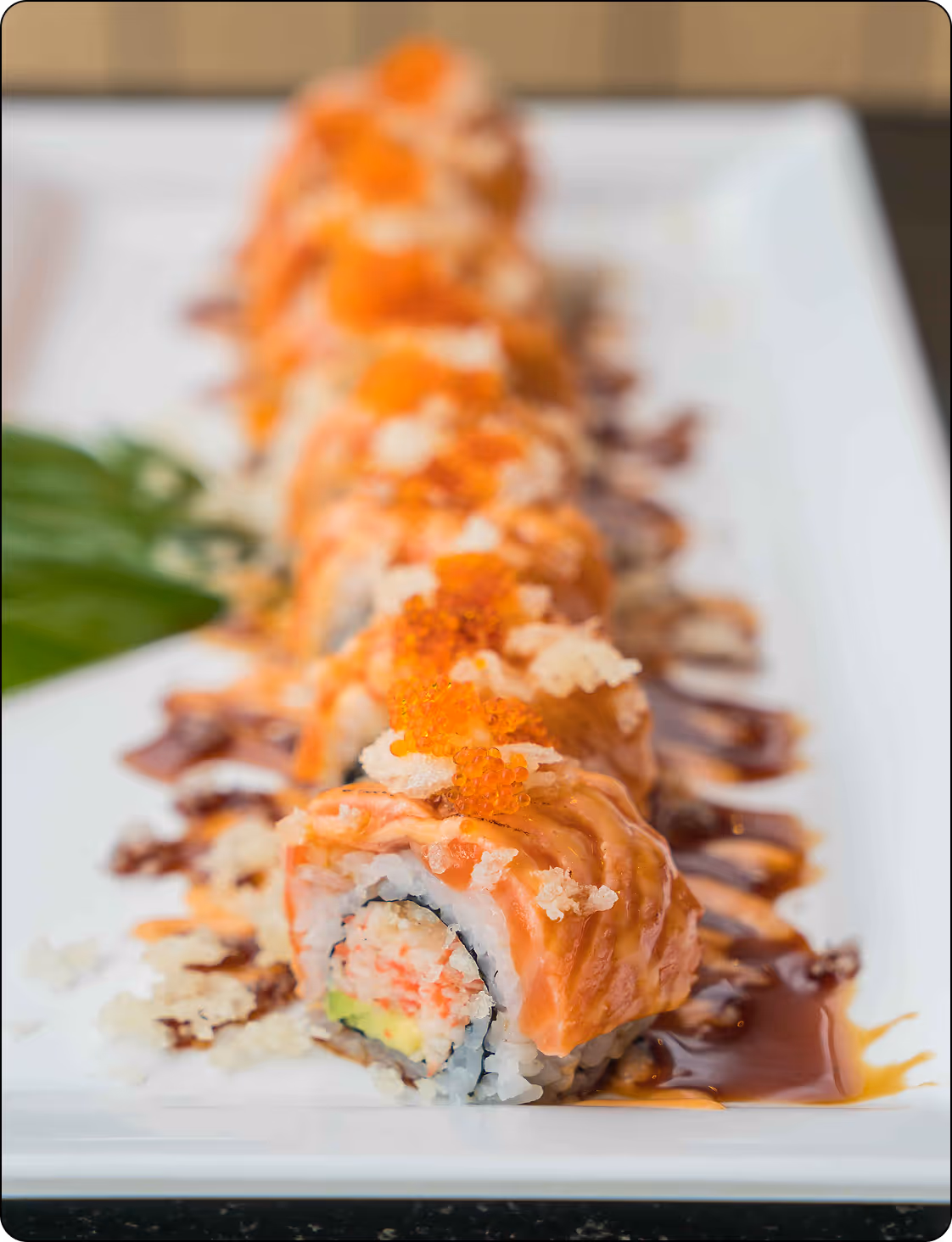 Close-up of sushi rolls topped with salmon, tempura flakes, and orange fish roe on a white plate with drizzle sauce.